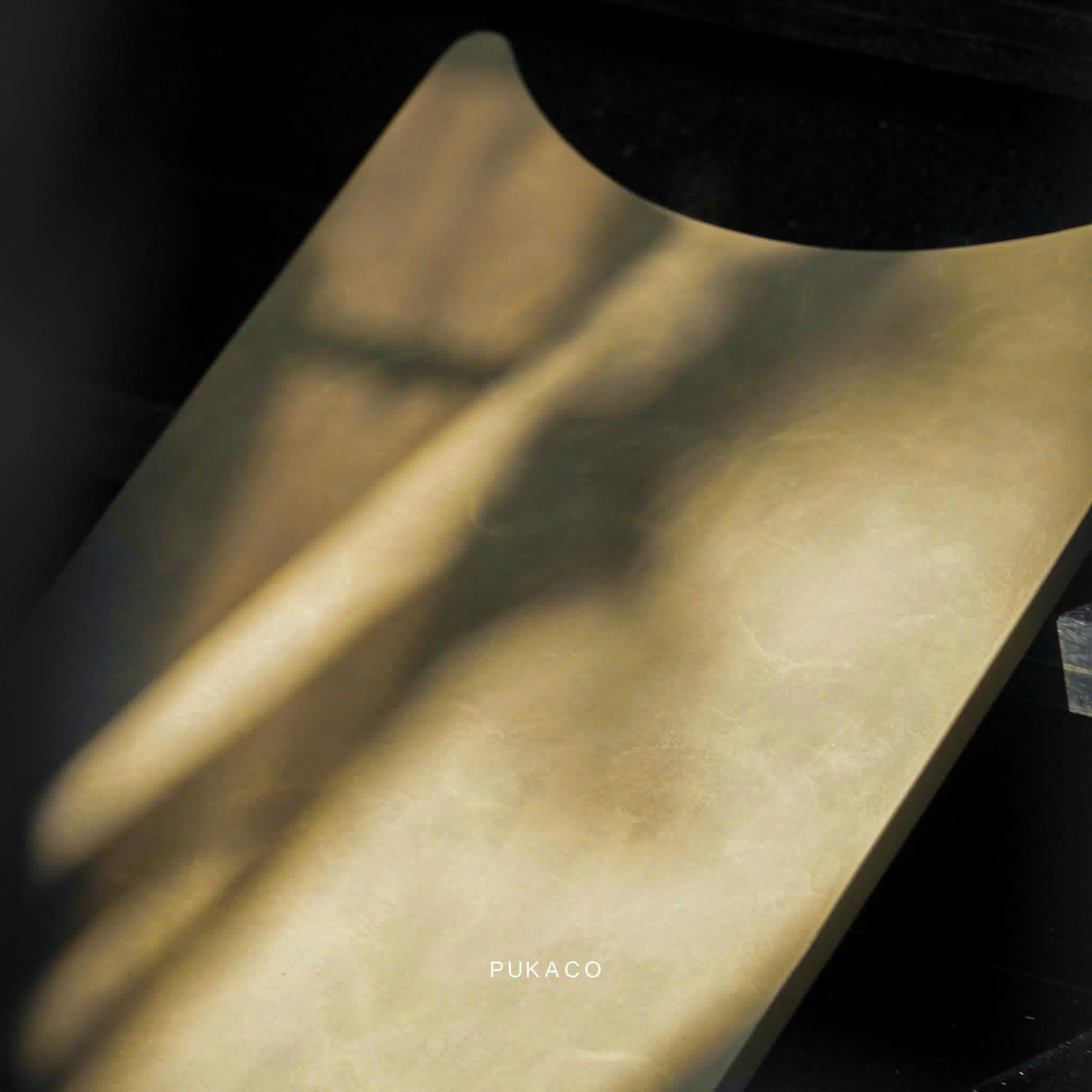 Original photograph of an ochre-toned sample with a curved cut edge, soft bands of natural light and shadow, and the original PUKACO watermark.