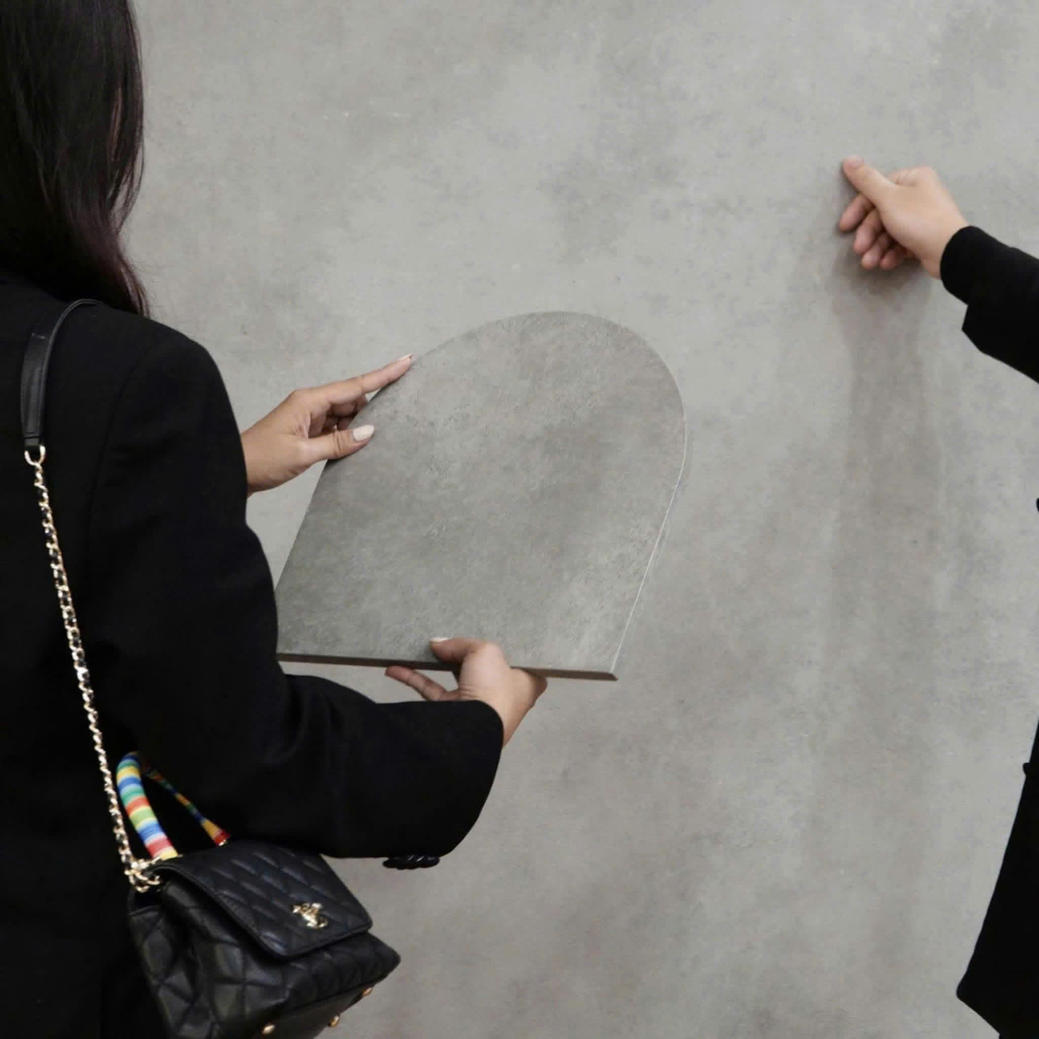 Detail from the original photograph of real hands holding a gray, round-topped physical finish sample against a larger wall surface.