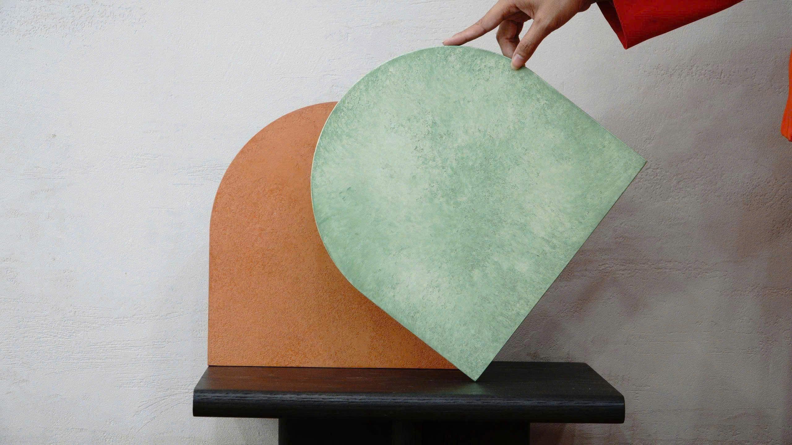 A hand holds a green finish sample beside a terracotta sample on a dark wood stand.