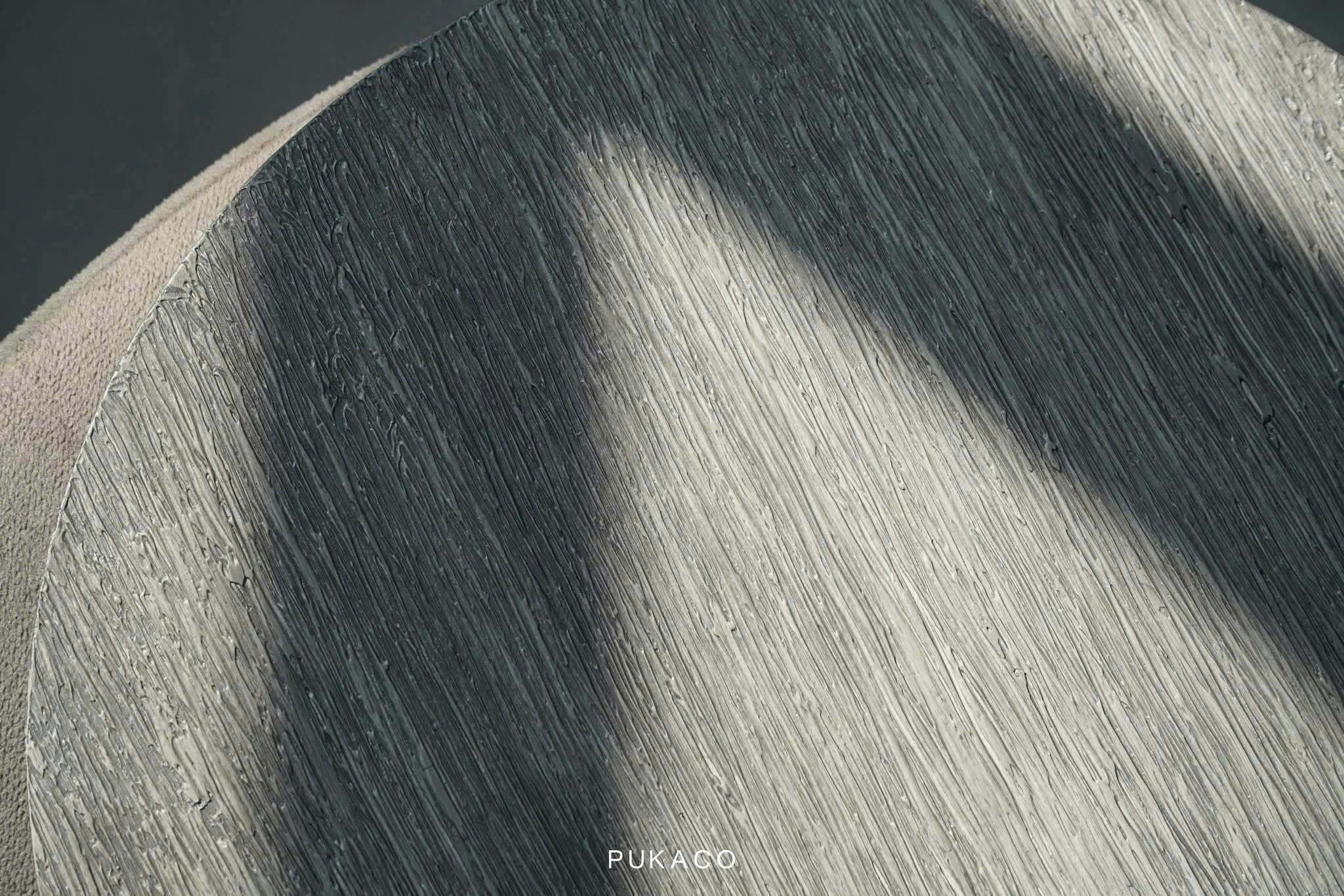 Raking light reveals fine hand-worked ridges across a gray PUKACO surface.