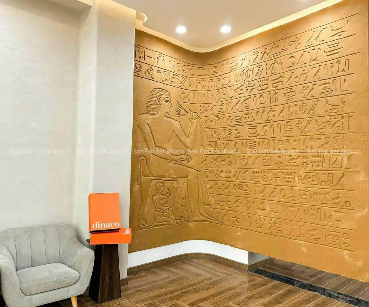 An installed curved ochre ECO CLAY wall with relief symbols beside a grey chair.