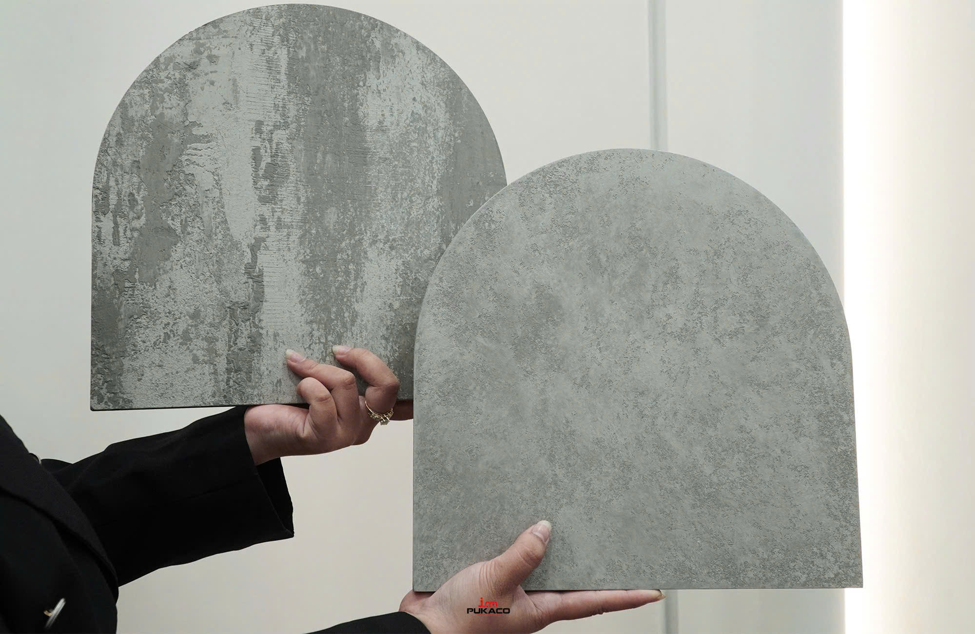 Hands hold two arched gray PUKACO finish samples with different surface textures.